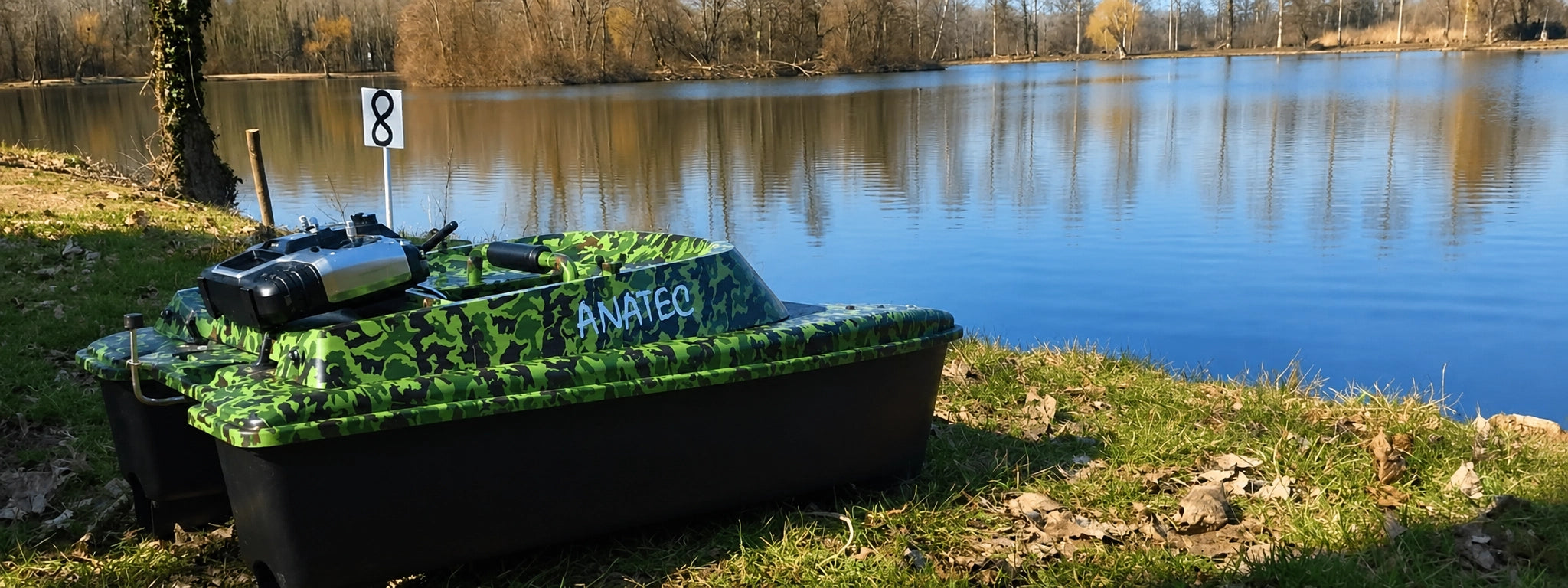 Bateau amorceur Anatec camouflage et sa télécommande posés sur la berge d'un étang