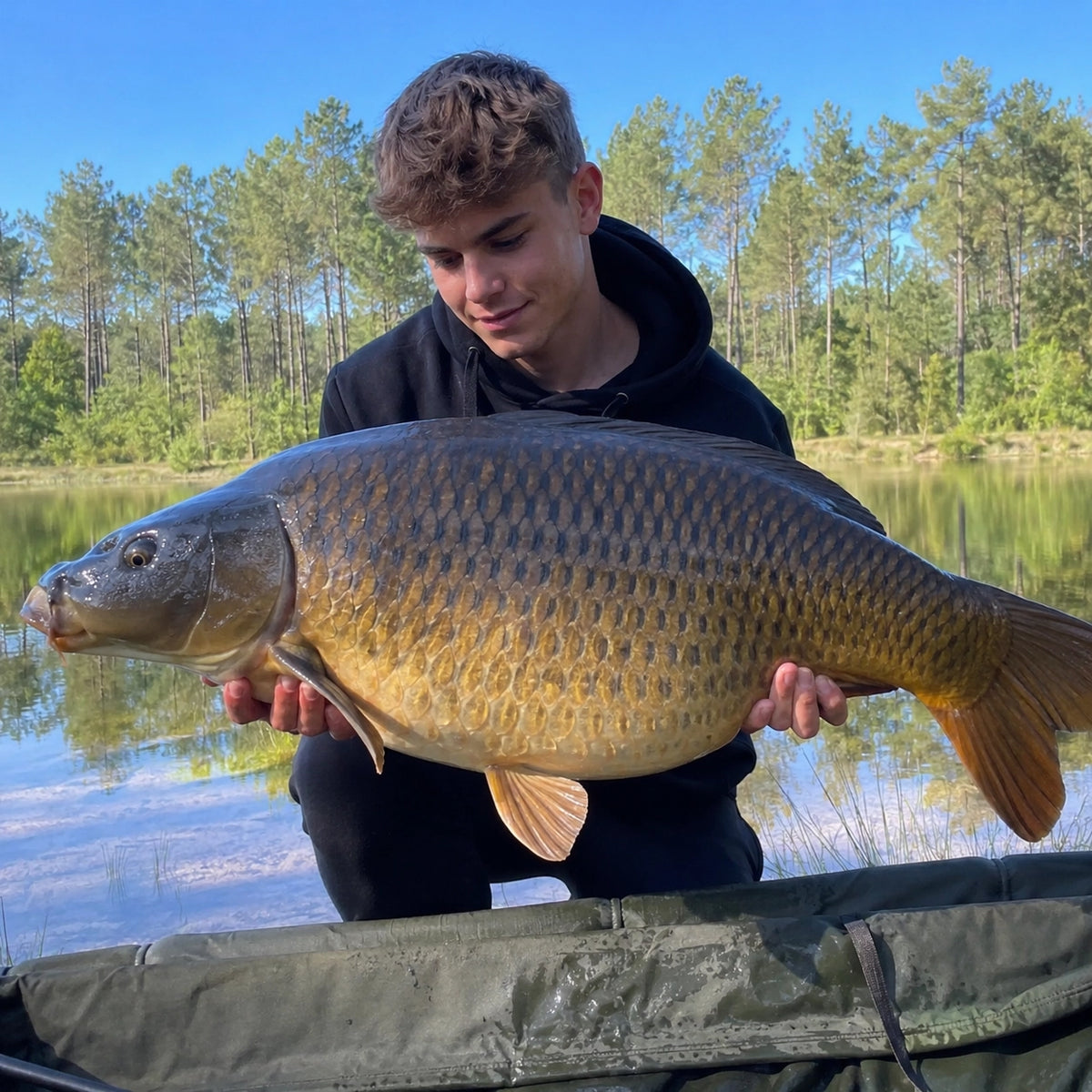 Jeune carpiste avec une carpe commune devant un lac bordé de pins landais