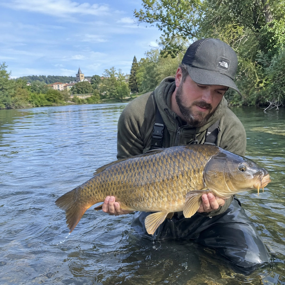 Pêcheur en waders tenant une carpe commune dans une rivière, un village d'Ariège en arrière-plan