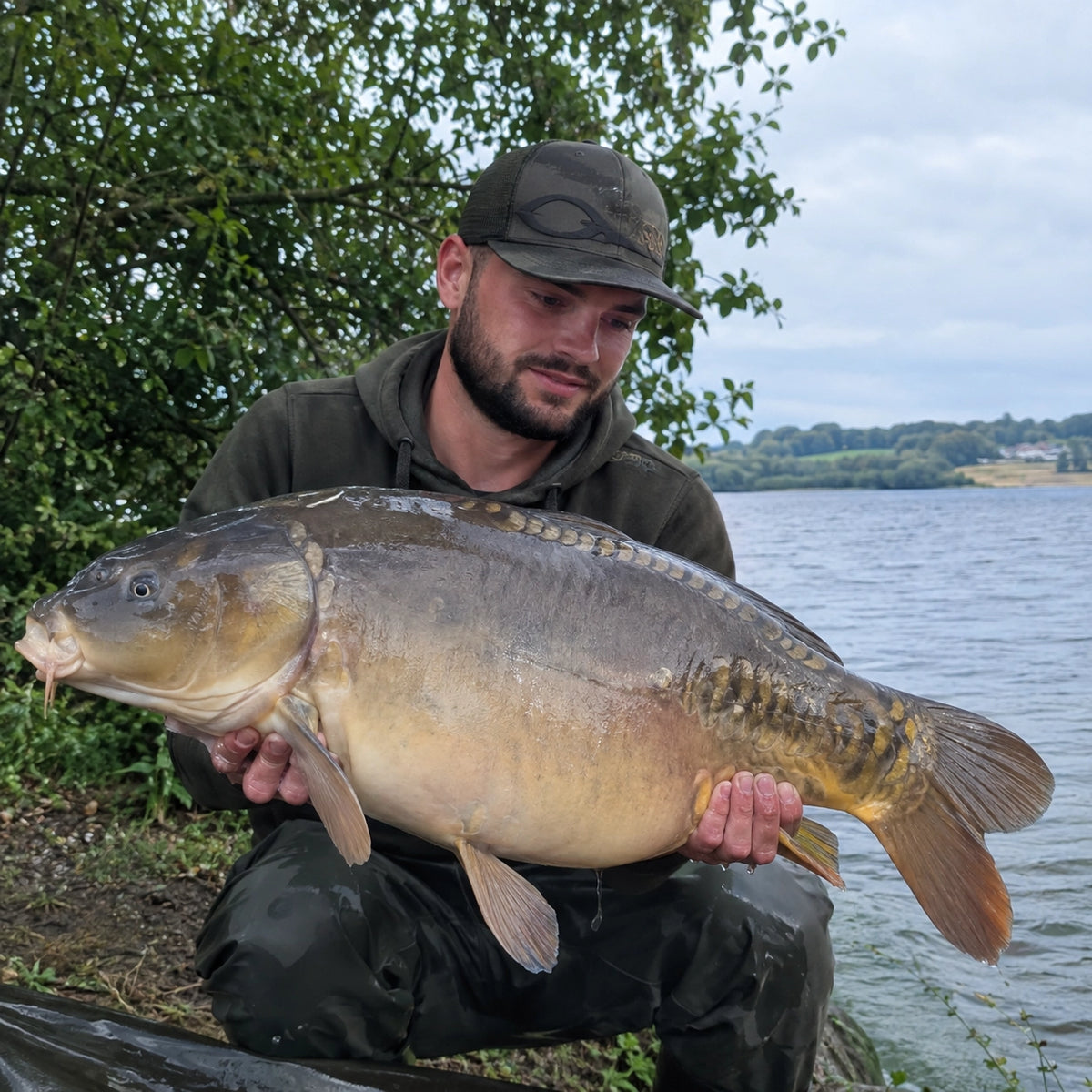 Carpiste en casquette présentant une carpe miroir au bord d'un lac, fruit d'une session bien préparée